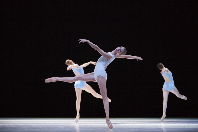 three dancers in white leotards balance on pointe. The woman in front looks out to the audience as she extends her back behind her
